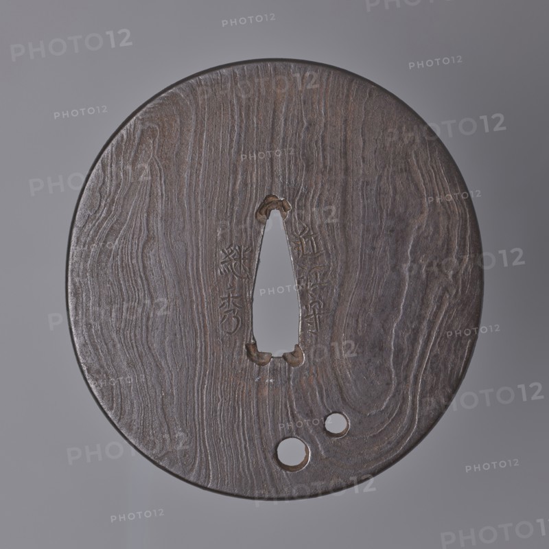 Crossguard tsuba, Tsuguhide. Oval tsuba tatemaru gata with a kaku mimi ko-niku type edge. Flat, forged from many layers, with two round holes udenuki ana, surface shaped like a tree trunk grain pattern mokume ji decoration. Signed. Note: the word kami lit. prince, lord is one of the titles used by tsuba masters and often included in signatures. Early 19th century, Japan, Ômi Eshu. Tsuba. Height 8.2 cm, width 7.7 cm, depth 0.4 cm., A round, flat object with a textured wood-like surface in shades of brown and dark brown sits against a neutral gray background; its center has a vertical, teardrop-shaped opening with rough, slightly darker edges and a few small carved notches nearby, and toward the lower edge there are a few circular holes; the surface shows flowing grain patterns and subtle variations of brown, dark brown, and hints of gray.