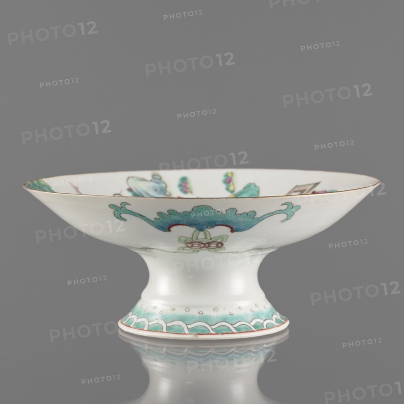Platter, gift; Paderewski, Ignacy Jan 1860-1941; Chinese art; Chinese; Qing dynasty 1644–1911, Tongzhi period 1862–1874; ceramics; colored porcelain; subject: go game, figural scenes, images of women, scholars, musical instruments; porcelain platter with white, transparent glaze; decoration painted with overglaze enamels and iron red, gilded edge; 1862–1874; China; vessel; height 8.5 cm; diameter 21 cm, A shallow pedestal bowl photographed against a plain gray background, with a white porcelain surface and decorative motifs in turquoise, green, red, and gold; the bowl has a flared rim with a thin gold edge, a concave interior showing small painted floral and ornamental elements in turquoise and green, and an outward scrolling turquoise motif on the exterior above a small stylized floral device in pale green and red; the pedestal foot is white with a band of turquoise wave-like decoration and small gold dots above a thin red line at the base, and the bowl and its foot are reflected fain...