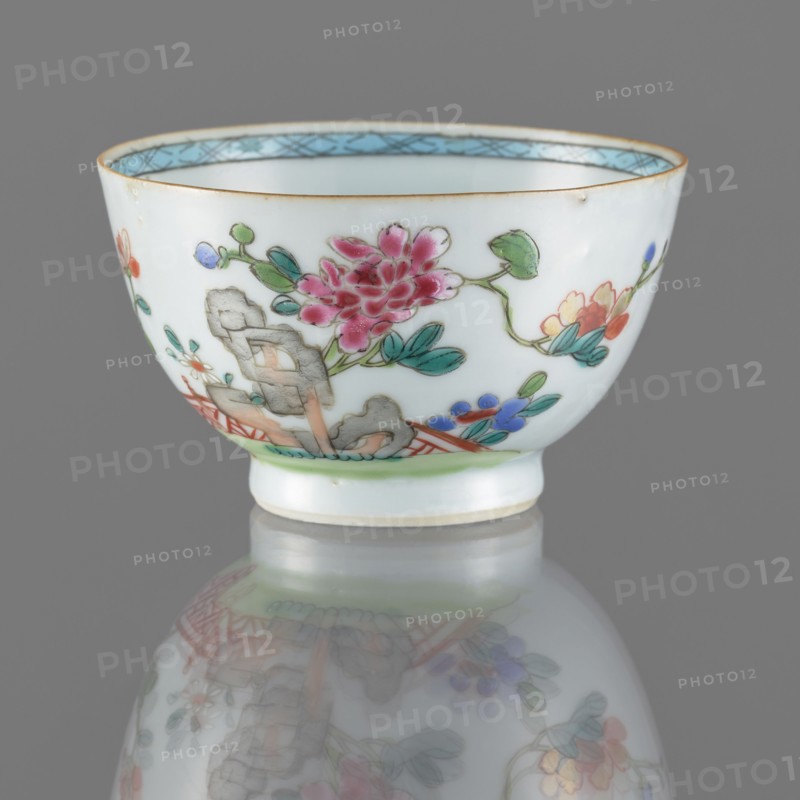 Bowl. Porcelain with white, transparent glaze with a bluish tint; decoration painted with overglaze enamel paints. Qing dynasty, 19th century. China. Spherical bowl form. Decor: flowers. Height 5.5 cm; diameter 8.8 cm., A white porcelain bowl with painted floral decoration sits on a reflective surface against a gray background, showing a large pink flower with green leaves, additional small flowers in shades of blue, yellow, orange, and purple, and a pale blue decorative band inside the rim; the bowl has a thin brown rim and a pale green line near the base, and its painted motifs and colors are mirrored softly in the reflection below.