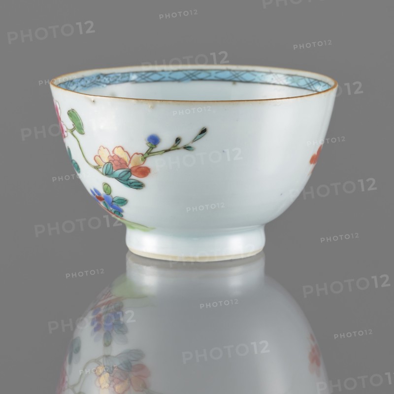 Bowl. Porcelain with white, transparent glaze with bluish tint; decoration painted with overglaze enamel paints. Spherical bowl form. 19th century Qing dynasty, 1644–1911. China. Colored porcelain. Height 5.5 cm; diameter 8.8 cm. Subject: flowers., A small white porcelain bowl with a rounded body and narrow foot, decorated on the exterior with a painted floral branch featuring petals and leaves in colors including green, blue, pink, orange, and yellow, with delicate dark green stems and small dark accents; the rim has a thin golden band and an inner border in light blue with a crosshatch pattern, and the bowl sits on a reflective gray surface that creates a blurred mirror image of the painted flowers.