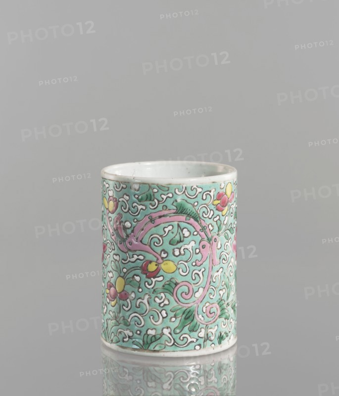 Brush vessel. Chinese; Qing dynasty 1644–1911. Porcelain with celadon glaze; decoration painted with overglaze enamel paints. Cylindrical brush vessel decorated with floral tendril and dragons. 19th century. Height 12.1 cm; diameter 9.6 cm; weight 0.5 kg., A cylindrical ceramic cup with a smooth white rim and base sits against a neutral gray background, its exterior covered in an intricate floral and scroll pattern in green, pink, yellow, white, and touches of red, with several leafy shapes and curling vine-like designs wrapping around the surface and a reflective surface beneath creating a faint mirrored image.