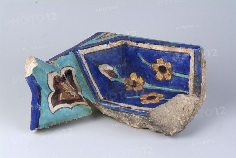 Fragment of a mukarnas, Islamic art, Persian culture, Timurid dynasty 1370–1526, Timur 1336–1405, architecture — mosques, fragments, architectural element, palmettes and floral ornament, glued, surface abrasions, end of the 14th century, Turkestan Kazakhstan, architectural element; height 21.4 cm, width 20.5 cm, depth 10.3 cm., A broken ceramic fragment with a glossy surface showing a deep blue interior decorated with floral motifs in gold, brown, turquoise, and black, bordered by a lighter blue edge and an off-white rim; the outer surface displays turquoise and cobalt blue areas with a stylized heart-shaped design in brown, cream, and black, and the piece rests on rough, unglazed beige-gray clay with some worn and chipped sections.
