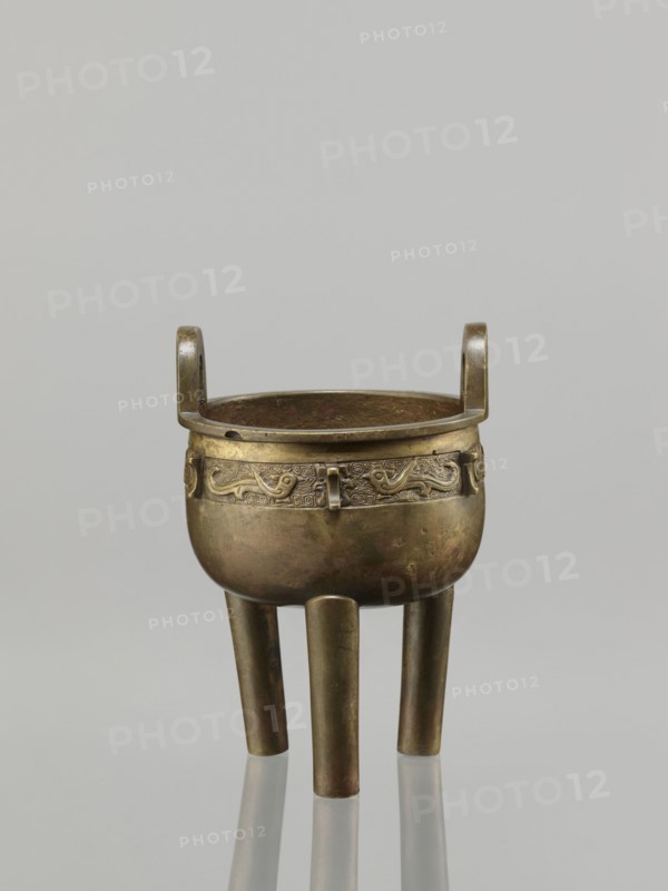 Ding incense burner, leiwen ornament, Chinese art, Ding vessel, Chinese culture, ceremonial/ritual vessel, birds decorative motif, meander ornament, Qing 1644–1911, 19th century, China, vessel, height 19.2 cm, diameter 13.6 cm, A metal tripod vessel with a rounded body and a pair of upward-curving loop handles sits against a plain light gray background; the vessel shows a metallic bronze and dark brown patina with areas of greenish tarnish and subtle reddish-brown spots, a decorative band with raised curvilinear motifs encircles the upper body, and three cylindrical legs extend downward, each reflecting slightly on the smooth surface beneath.