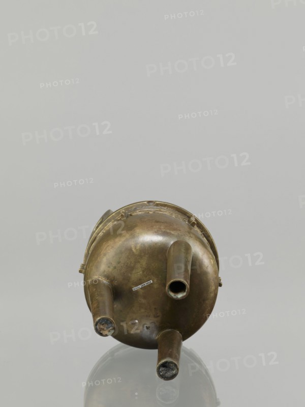 Ding incense burner, leiwen ornament, Chinese art, Ding vessel, Chinese culture, ceremonial/ritual vessel, decorative birds motif, meander ornament, Qing dynasty 1644–1911, 19th century, China, vessel, height 19.2 cm, diameter 13.6 cm, A metallic, round, aged object with a patina of brownish bronze and greenish tarnish sits upright on a reflective surface, showing a rounded base with three tubular protrusions extending outward; the metal has darker and lighter patches and some worn areas, and the reflection and neutral background display shades of gray and muted silver.
