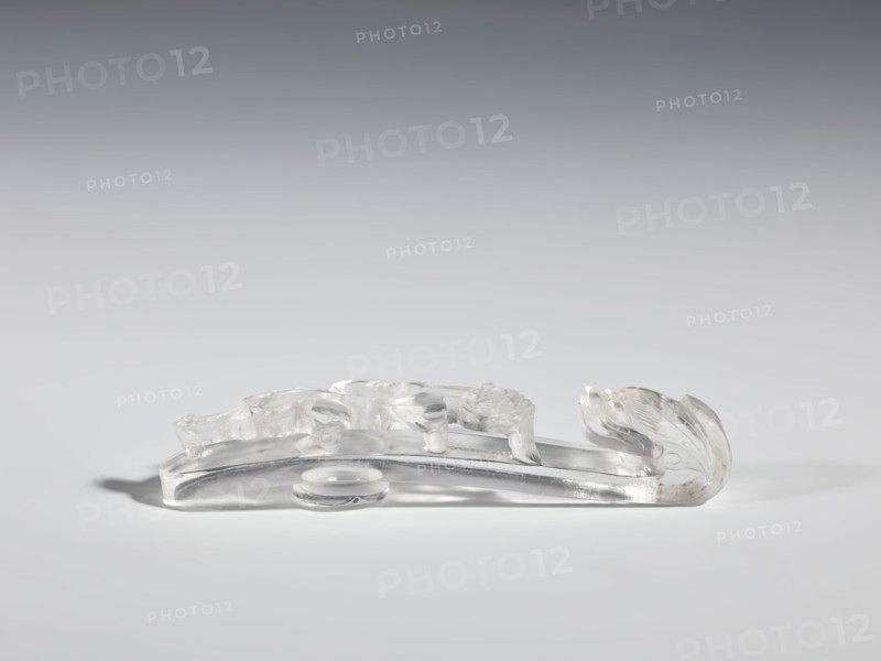 Belt clasp, Chinese; Qing 1644–1911; 17th–19th century. Material: rock crystal with inclusions. Subject/iconography: lizard-dragon chilong. Description: full-plastic carved head; shank with deep relief, openwork, and engraving. Function: belt buckle for men's clothing. Dimensions: height 2.0 cm, width 9.8 cm, depth 2.0 cm., A translucent carved object rests on a smooth surface, showing clear and frosted areas with glossy highlights; its elongated shape curves upward at one end into a rounded, leaflike form with carved grooves, while the opposite side features low-relief rounded motifs and gentle undulations; the piece casts a soft shadow and the visible colors are clear, white, gray, and subtle silvery tones.