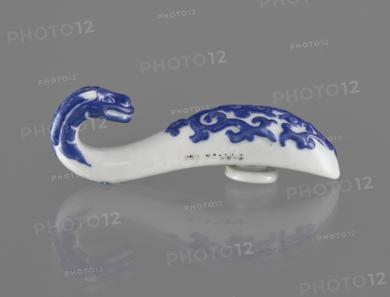 Belt clasp., Chinese, 19th century. Shaft: stylized lizard-dragon, bats on the underside. Navy blue overglaze enamel paint on colored porcelain. Function: belt buckle. Dimensions: height 3.1 cm, width 9.3 cm, depth 2.65 cm., A small curved ceramic object shaped like an animal with a curled neck and head, resting on a flat reflective surface, is shown against a plain gray background; the object is primarily white with areas of deep blue forming a stylized pattern on its back and blue detailing on the head and neck, and its reflection in the surface mirrors the white and blue colors.