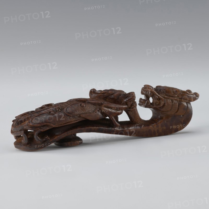 Belt clasp, Chinese; dragons iconography; Qing 1644–1911; 19th century; men's clothing ornament — belts. Shaft: two fighting lizard-dragons. Full-sculpture carved head; openwork shank. Materials/technique: carved. Dimensions: height 3.0 cm, width 12.8 cm, depth 2.8 cm., A carved wooden object in dark brown tones shaped like a stylized creature with an elongated curved body, a pronounced open-mouthed head with visible teeth, and intricately carved flowing details along its back and sides, resting on a light neutral surface against a soft gradient background of pale gray to muted bluish-gray.