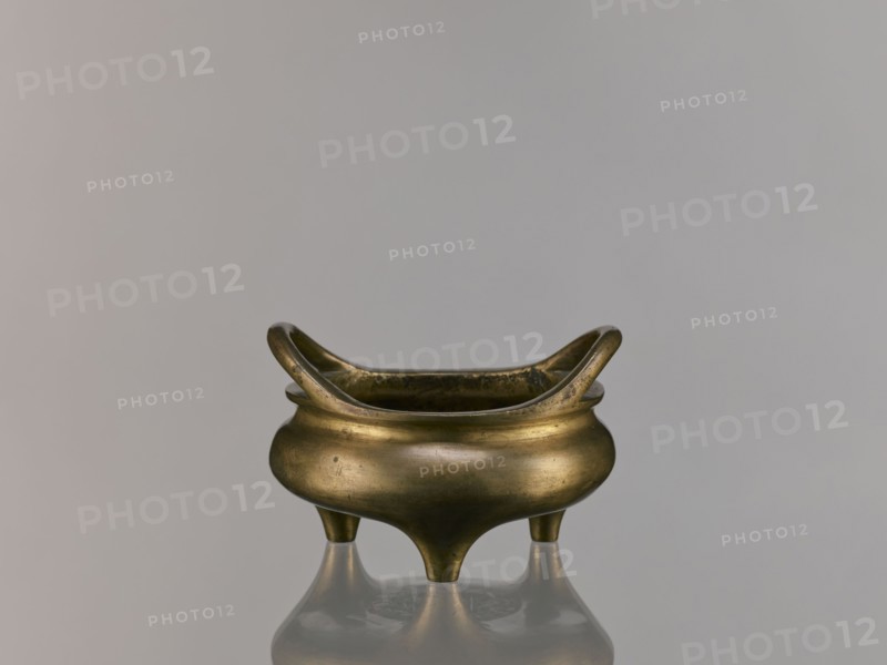Li-type incense burner, Li-type vessel, Qing dynasty 1644–1911, 18th century China; ceramic vessel/ceremonial ritual incense burner with fantastical beasts; backdated signatures; height 7.5 cm, width 12.1 cm., A small metal vessel with a rounded body and a curved, flaring rim sits centered on a reflective surface; the vessel has a warm brass color with darker patina spots and subtle surface marks, and it stands on three short tapered legs that cast reflections below, set against a smooth pale gray background.
