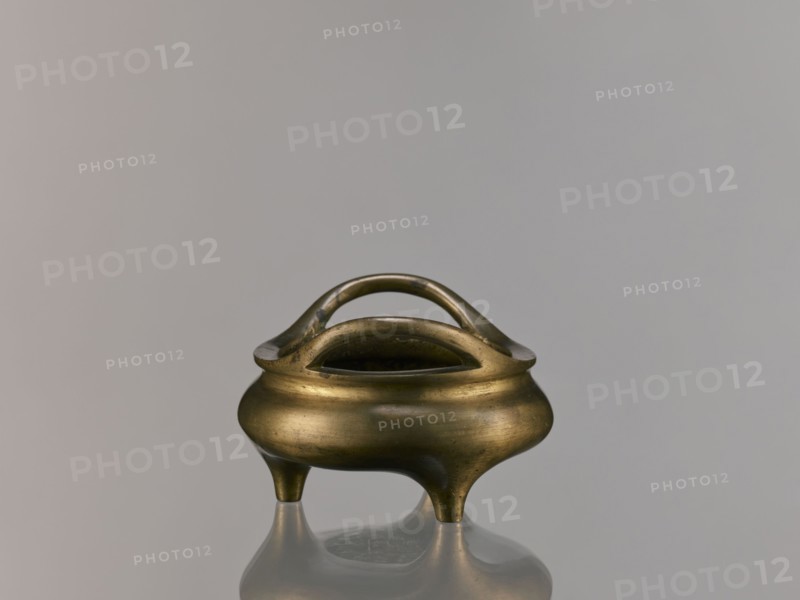 Li-type incense burner, Li-type vessel, Qing dynasty 1644–1911, 18th century, China. Bronze vessel in Li form used as an incense burner, decorated with fantastical beasts; backdated signatures; height 7.5 cm, width 12.1 cm., A small metallic vessel with a rounded body and a warm brass tone sits on a reflective surface, supported by three short tapered legs and topped by a smooth curved double-handle arch; the vessel shows subtle surface variations and patina in shades of brass, gold, and darker brown, while the background is a soft neutral gray.