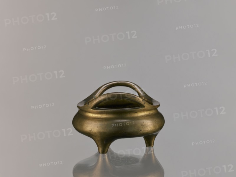 Li-type incense burner, Chinese art, Chinese culture, ceremonial vessel, Li-type vessel, Qing 1644–1911, fantastical beasts, ritual vessel, Qing style, backdated signatures, 18th century, China, vessel, incense burner, height 7.5 cm, width 12.1 cm, A small rounded metal vessel with a warm brass color sits on a reflective surface, showing a soft reflection below; the vessel has a bulbous body tapering into three short rounded legs and features a double-arched handle that rises above an open central rim, with the metal surface displaying subtle darker spots and a slightly aged patina against a plain light gray background.