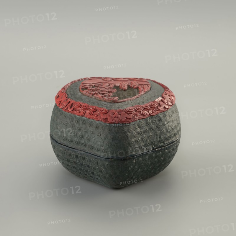 Box with a lid, Chinese, Qing 1644–1911, 19th century. Two-tone lacquer with carved decoration depicting figurative scenes: peaches, flowers, bats, pines, rocks, clouds, kikko pattern. Container, height 7.3 cm, width 10.6 cm, depth 10.5 cm., A rounded, textured container with a muted green surface and a decorative red band around its top edge sits on a neutral light-gray background; the container has a patterned, slightly rough exterior and a lid topped with an ornate red raised design, showing a contrast between the green body and the red decorative elements.