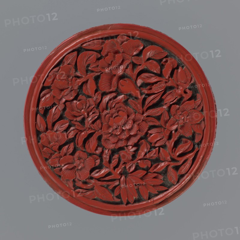 Box with lid, Chinese, Qing dynasty 1644–1911, 19th century. Two-tone lacquer red and black; black lacquer inside. Decorated with floral ornament: peonies, bamboos, peaches iconography, kikko geometric pattern. Function: container. Dimensions: base height 2.1 cm, diameter 8.8 cm; lid height 1.8 cm, diameter 8.8 cm; overall box height 3.8 cm, diameter 8.9 cm., A round carved object with a raised floral relief occupying the center, featuring a group of intricately carved flowers and many leaves arranged densely; the carved elements are predominantly red with darker recesses between petals and leaves creating contrast, and the piece is framed by a red circular rim set against a gray background.