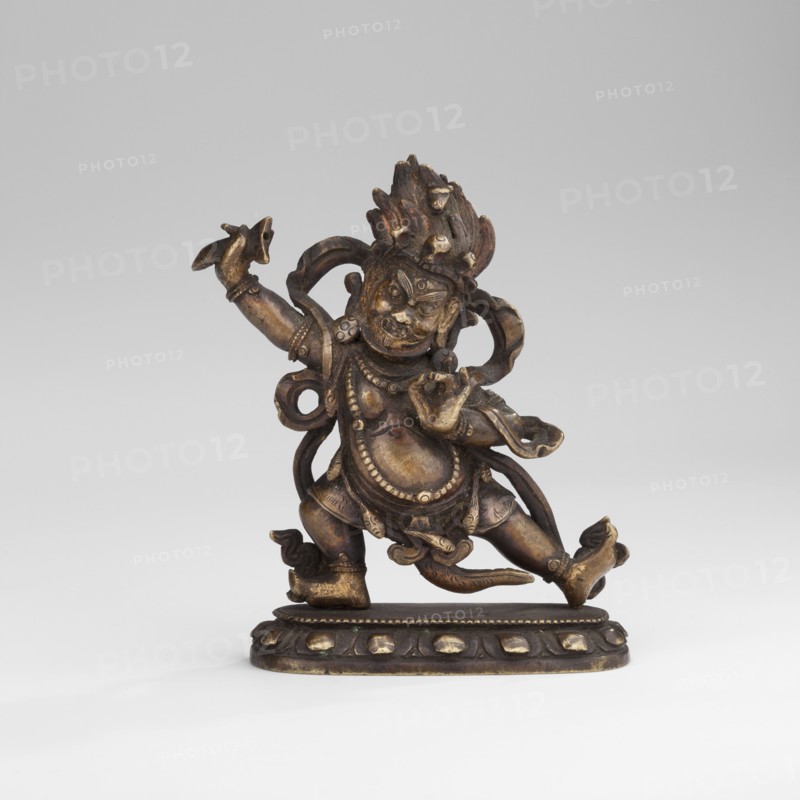 Hayagriva, unknown sculptor, Buddhist art, Himalayan art, Tibetan Buddhist art, Himalayan culture, dharmapala iconography, 2nd half of the 18th century, Tibet, sculpture, figurine; height 7.0 cm, width 6.1 cm, depth 2.4 cm, A small bronze figure with a dark brown and golden patina stands on an oval base; the figure has a dynamic, dancing pose with one leg raised and one arm extended upward, wearing ornate jewelry and draped cloth elements, and a headdress with textured, flame-like details. The surface shows areas of worn gold highlighting on raised features, with darker brown recesses throughout. The base is dark brown with golden accents on rounded decorative elements, and the background is a smooth light gray.