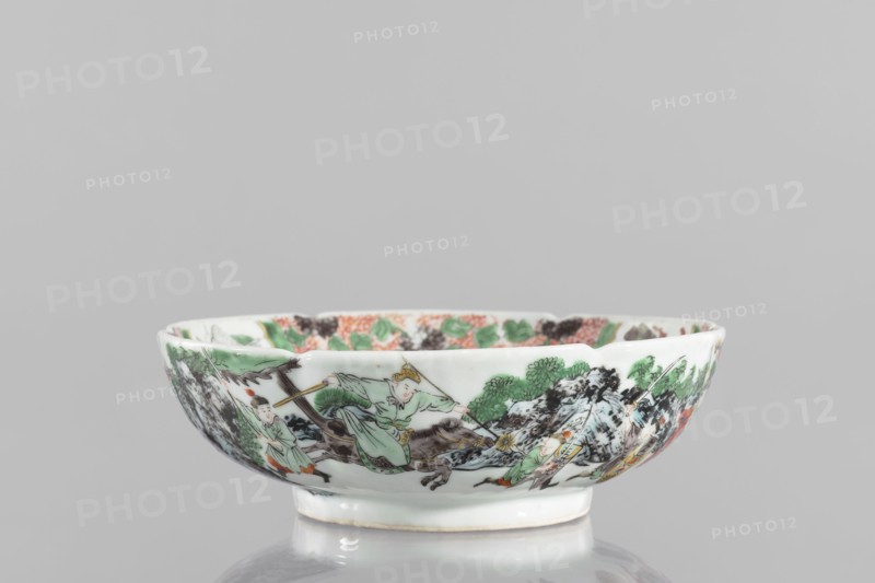Bowl., Chinese; Qing dynasty 1644–1911. 19th century. Porcelain body with white, transparent glaze; decoration painted with overglaze enamel paints and iron red depicting figural and knightly scenes. Six-leafed belly form. Height 6.5 cm; diameter 20.0 cm; weight 0.75 kg., A shallow porcelain bowl with a scalloped rim sits on a reflective surface; the bowl is primarily white and decorated around its exterior with painted scenes in green, black, brown, orange, and touches of yellow, showing a group of human figures in flowing garments, stylized trees and foliage, and rocky landscape elements; the interior rim also has painted floral and foliage motifs in green, orange, and black.