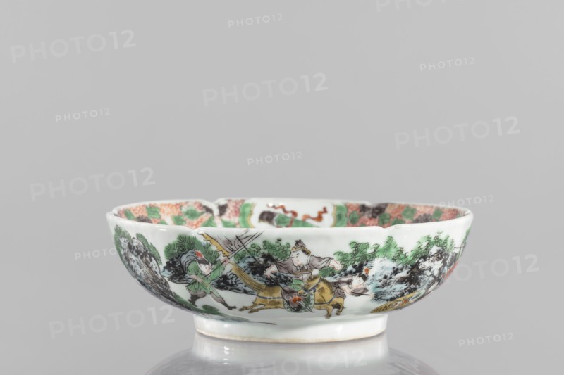 Bowl. Porcelain body with white, transparent glaze; decoration painted with overglaze enamel paints and iron red. Colored porcelain depicting figural scenes and knightly scenes; six-leafed belly. Qing dynasty 1644–1911, 19th century, China. Height 6.5 cm; diameter 20.0 cm; weight 0.75 kg.,  noted. Chinese., A shallow porcelain bowl with a white base color sits on a reflective surface, decorated around its exterior with a painted scene featuring a group of human figures and animals amid stylized landscape elements. The painted colors include green, black, brown, yellow, beige, pink, and hints of red and orange. The bowl has a slightly flared rim and a small foot, and the interior rim shows patterned decoration in similar colors. The surface appears glossy and smooth, and the background behind the bowl is a uniform light gray.