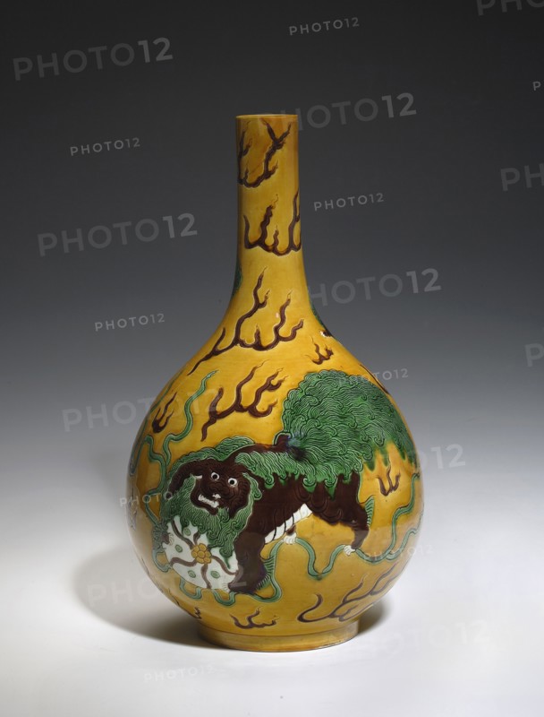 Onion-shaped vase, lion iconography, ceramics, Chinese art, Chinese culture, vases, Qianlong 1736–1795 style, pre-dated signatures. Vase with a narrow, long neck. Decoration on a yellow background: five lions—one larger with a ball and four smaller ones among flames. Yellow glaze, white in parts of the decoration; engraved decoration, painted with green and brown enamel paints. 19th century, China. Vessel. Height 32.0 cm; diameter 18.5 cm., A bulbous ceramic vessel with a long narrow neck sits against a dark gray background, the vessel painted a vivid yellow with flowing brown flame-like motifs; a stylized creature with a dark brown body and a textured green mane is depicted on the rounded body, standing over a pale circular object and surrounded by sinuous green tendrils, with highlights of white and small areas of lighter yellow on the base and shadowed white surface beneath.