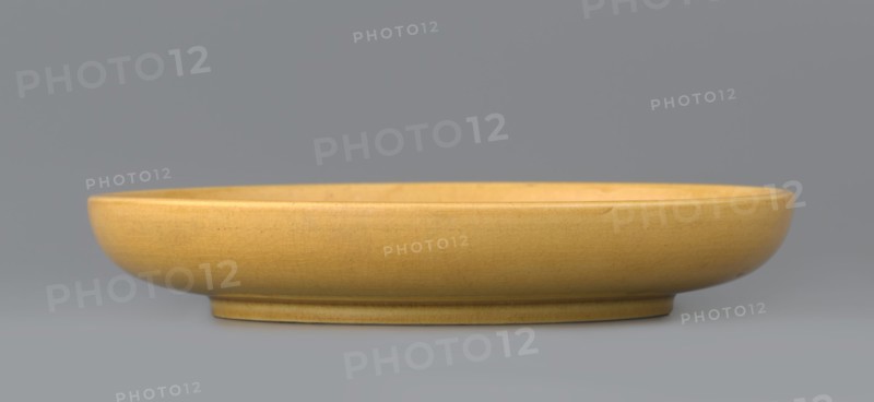 Plate, pheasants; ceramics; Chinese art; Chinese culture; Qing dynasty 1644–1911, late 19th century; monochromatic yellow glaze, finely crackled; engraved decoration; the mirror depicts a pheasant; vessel plate; height 2.5 cm, diameter 14.6 cm., A shallow round dish with a smooth rim and a low foot, shown in profile against a plain background; the dish is a warm yellow color with subtle crackle texture and small darker specks, and the background and surface beneath the dish are shades of gray.