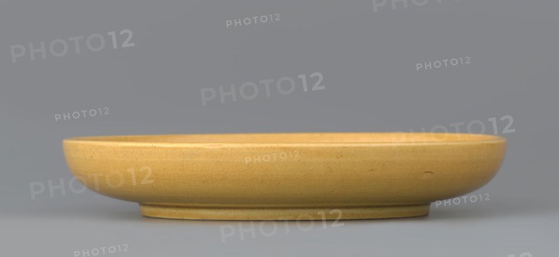 Plate, pheasants, ceramics, Chinese art, Chinese culture, Qing style, plates, Qing 1644–1911, monochromatic decoration, yellow; the mirror depicts a pheasant. Yellow glaze, finely crackled, engraved decoration, late 19th century, China, vessel, height 2.5 cm, diameter 14.6 cm, A shallow round ceramic dish with a smooth curved rim and a slightly raised foot, shown from the side against a neutral background; the dish is a warm yellow color with subtle crackle texture and small darker speckles, and the background and surface beneath the dish appear in shades of gray.
