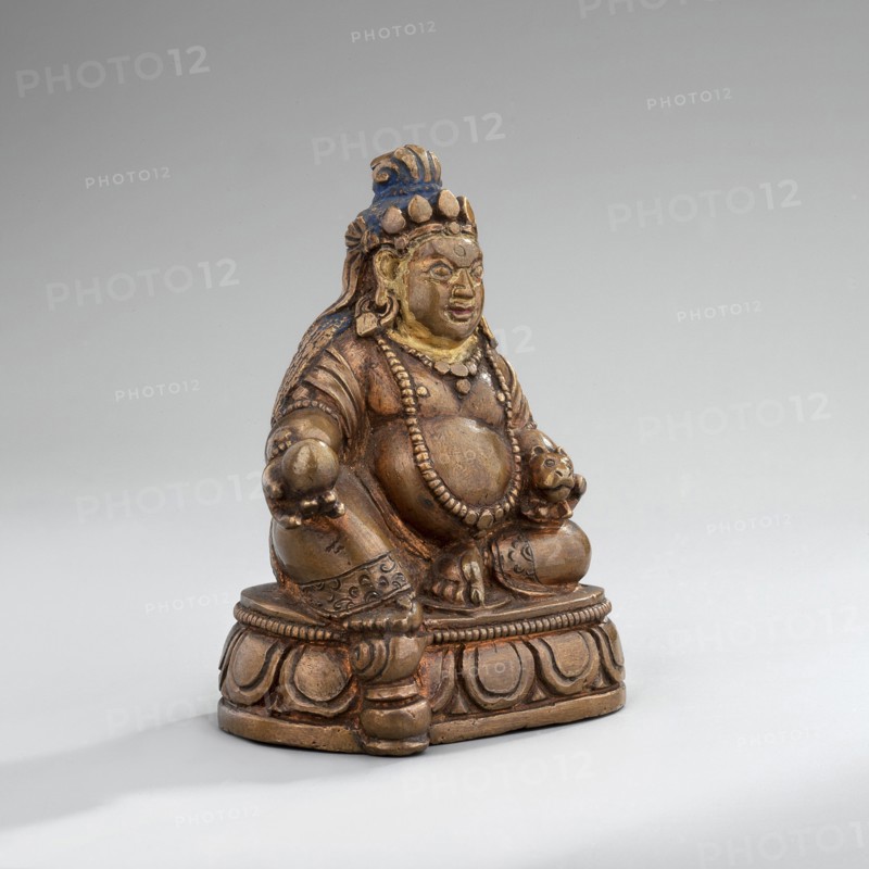 Jambhala, Himalayan culture, Tibetan Buddhist art, Himalayan art, dharmapala iconography, lotus throne iconography, cast copper, gilded face later; polychrome lips, eyes, and hair, mid-19th century, Mongolia, sculpture, figurine, height 6.7 cm, width 5.3 cm, depth 4.0 cm, A small seated figurine made of a brownish metal with areas of golden and bluish paint sits on a rounded ornate base; the figure has a rounded belly, beaded necklaces, and detailed carved garments and jewelry, with a headdress that shows blue and gold, a serene facial expression, and hands resting on the knees while one hand holds a small rounded object; the base and figure show carved decorative patterns and a worn, aged surface with visible highlights of gold, brown, and blue against a pale gray background.