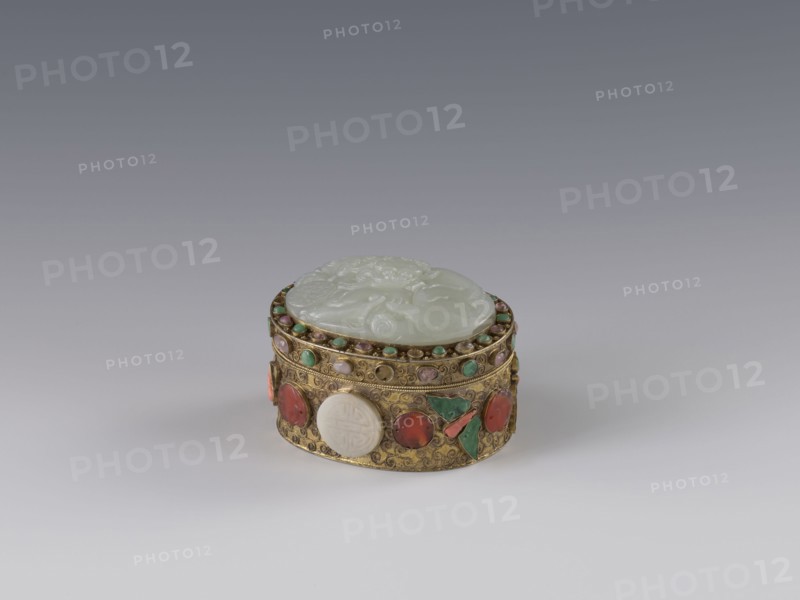 Box with lid. Chinese; Qing dynasty 1644–1911, late 19th century. Lid inlaid with a jadeite plaque carved in bas-relief showing a fo lion playing with a ball. Outer walls decorated with motifs of butterflies, bats, and the shou longevity symbol made of semi-precious stones in settings. Background shows a plant twig with filigree. Material: semi-precious stones and jadeite. Function: vessel/container. Dimensions: height 5.0 cm; width 6.6 cm; depth 8.2 cm; weight 0.17 kg., A small oval decorative box sits on a neutral gradient surface, with a pale translucent carved lid that is smooth and slightly raised, showing shallow carved motifs. The body of the box is metallic gold with intricate engraved patterns and raised bezel settings holding a group of round and oval stones in colors including red, green, turquoise, pale pink, and white, with a few triangular green stone pieces set decoratively. The gold metal has detailed filigree work and a band of small round turquoise and pink beads alon...