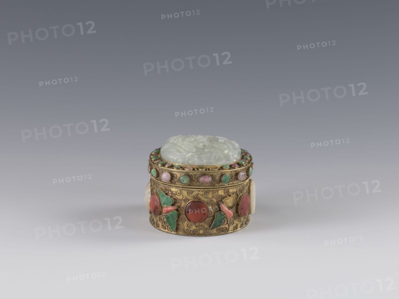 Box with lid;, Chinese, Qing dynasty 1644–1911, late 19th century. The lid is inlaid with a jadeite plaque with a bas-relief image of a fo lion playing with a ball. The outer wall features motifs of butterflies, bats, and the shou symbol of longevity made of semi-precious stones set in settings. Background: plant twig with filigree. Vessel/container. Height 5.0 cm; width 6.6 cm; depth 8.2 cm; weight 0.17 kg., A small round decorative box sits centered on a smooth gradient background, with a pale green carved lid resting on an ornate golden body; the gold surface is decorated with a pattern of swirls and set with several small round stones in shades of green and pink and a few larger rounded cabochons in red and coral tones, along with green leaf-shaped inlays and pale cream circular side accents.