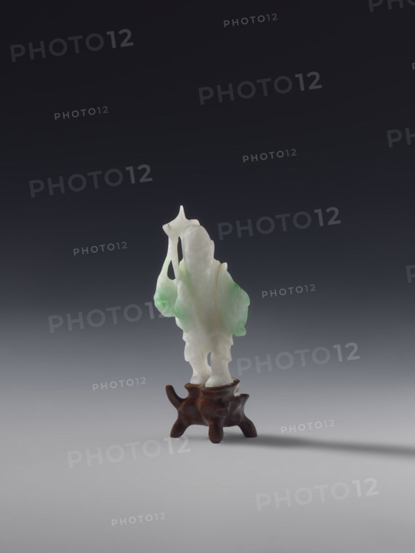 Fisherman with a Basket and a Fish on a Rod, unknown sculptor, fish, fishermen, Chinese art, Chinese culture, Qing 1644–1911, White jade with green spots, solid sculpture, engraved, 19th century, China, figurine, sculpture, height 7.2 cm, A small carved figurine made of a translucent white and pale green material stands on a dark brown wooden base; the carving has smooth, flowing contours with irregular, rounded forms and thin protrusions, and the base is organic-shaped with curved legs and a polished surface; the background is a gradient from dark charcoal at the top to light gray at the bottom, and a soft shadow extends from the base across the gray surface.