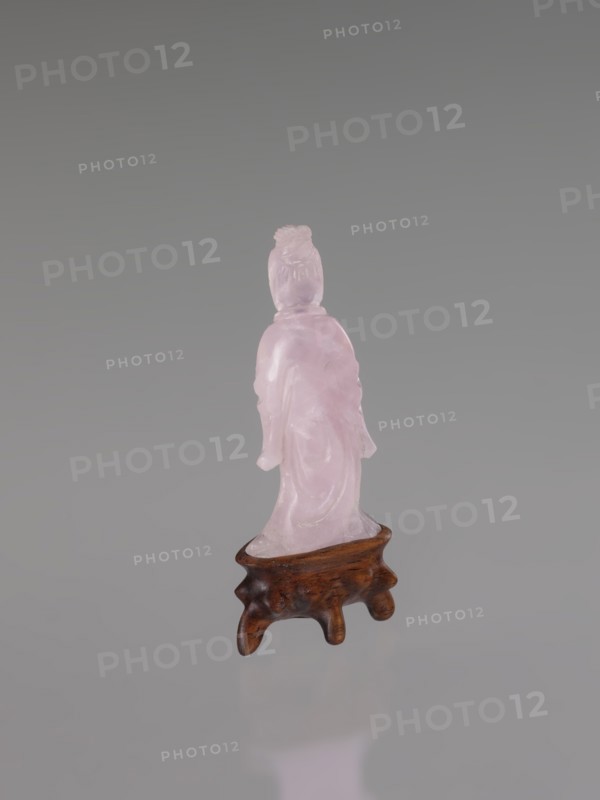 Guanyin on a base, Chinese art, Chinese culture, Qing 1644–1911, Chinese Buddhism, goddess, figurine carved in rose quartz, full sculpture, engraving, 19th century, China, sculpture, height 6 cm, A translucent pink carved figure standing on a small wooden stand with a polished brown finish; the figure appears to wear draped clothing and has a textured hairstyle, and the background is a smooth gradient of gray with reflections of the pink and brown tones visible on the surface below.