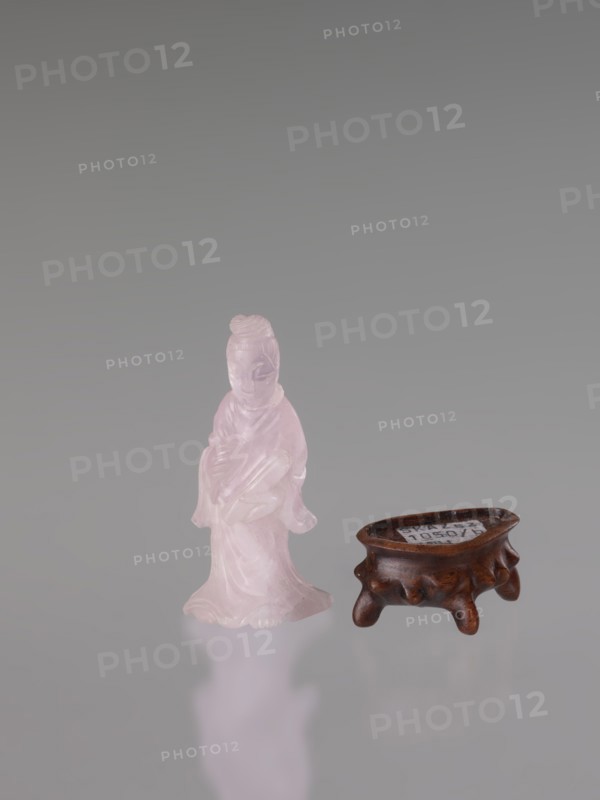 Guanyin on a base; figurine carved in rose quartz; full sculpture with engraving; Chinese; Qing dynasty 1644–1911; Chinese Buddhism; goddess; 19th century; sculptor, height 6 cm., A small translucent pink figurine of a robed person standing beside a low dark brown wooden stand with short rounded legs; the background is a smooth gradient of gray and the surface beneath reflects faintly with soft pink and gray tones.