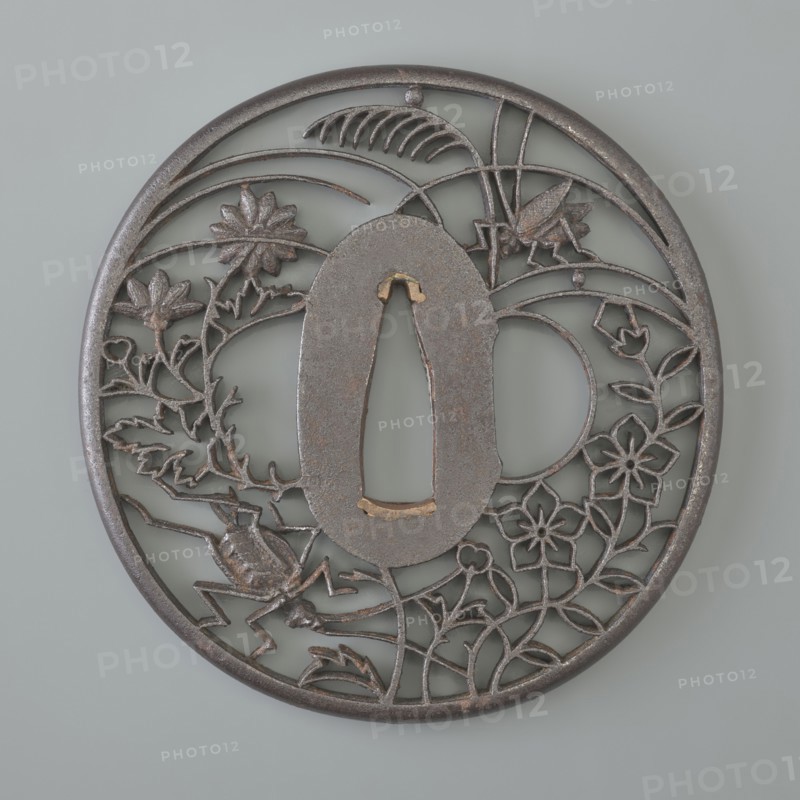 Crossguard tsuba, Akasaka role classification of creator, school author; Japanese; Edo period 1603–1868, 18th/19th century. Tsuba round maru gata with a rounded edge maru mimi; seppa dai oval; sekigane copper. Decorated with openwork ji sukashi decoration, partly positive and partly negative, depicting two crickets among chrysanthemums, flowers, and grasses. Not signed. Function: sword crossguard katana. Diameter 7.2 cm; thickness 0.5 cm., A round metal object with a dark gray to brownish patina sits against a pale gray background; the piece has an openwork design featuring a central oval cutout framed by a ring of thin metal, with floral motifs and leafy vines woven throughout the circular field, a few stylized flowers and several curved leaf shapes, and a couple of insect-like forms rendered in the same dark metallic tones.