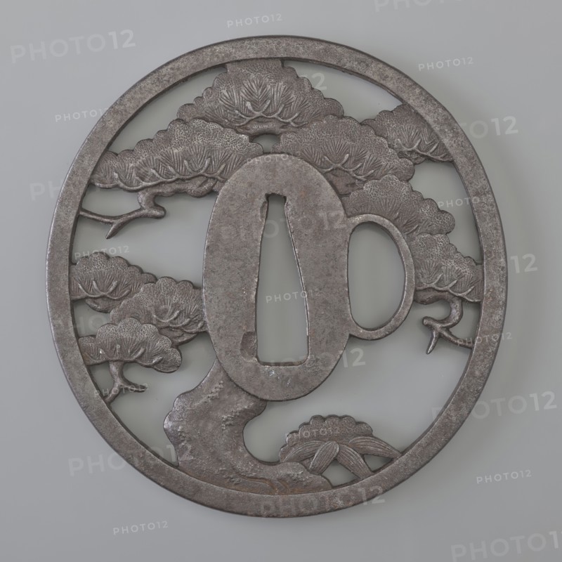 Crossguard tsuba, Sunagawa, Masayoshi, / personal slogans / artist, Sunagawa,  school author; Japanese; Edo period 1st half of the 19th century; iron tsuba, round tatemaru gata with one side hole katahitsu ana and massive rectangular edge kaku mimi. Openwork positive composition depicting a spreading pine tree with bamboo growing from its thick trunk, marked with a few leaves; seppa dai oval; kozuka hitsu ana woven into the decorative motif but clearly visible. Signed. Height 7.75 cm, width 7.4 cm, thickness 0.5 cm., A round metal object with a dark gray, weathered surface features an openwork design of stylized tree forms and cloud-like foliage; a central oval element with an open vertical slot and a rounded side opening sits over a textured trunk and branches, and the background shows a few layered cloud-tree shapes and curved limbs. The visible colors are dark gray, lighter gray, and pale gray.