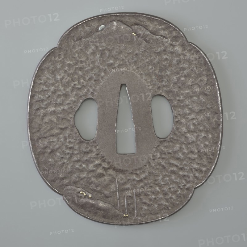 Crossguard tsuba, attributed to Kaneie craftsman/school. Tsuba in the shape of a quatrefoil mokkô gata with a slightly raised edge uchi kaeshi and ryôhitsu ana, textured surface ishime ji. Decorated in low relief hirabori with a landscape motif: a fisherman sitting on the water's edge under a cliff, moon emerging from behind distant hills; motifs include rocks, mokkô, bamboos, trees, mountains. Signed. 18th century Edo period, 1603–1868, Japan — Fushimi, Yamashiro. Materials/genre: Japanese sword crossguard tsuba. Dimensions: height 8.5 cm; width 7.8 cm; depth 0.4 cm., A roughly oval, slightly scalloped metal object with a textured, dimpled surface in shades of dark gray and muted silver sits against a pale gray background; it has three central openings with smooth edges and a vertical central cutout flanked by two rounded cutouts, and small brighter silver highlights along the rim and inside the cutouts; a few tiny gold-colored flecks are visible near one scalloped edge and another sm...