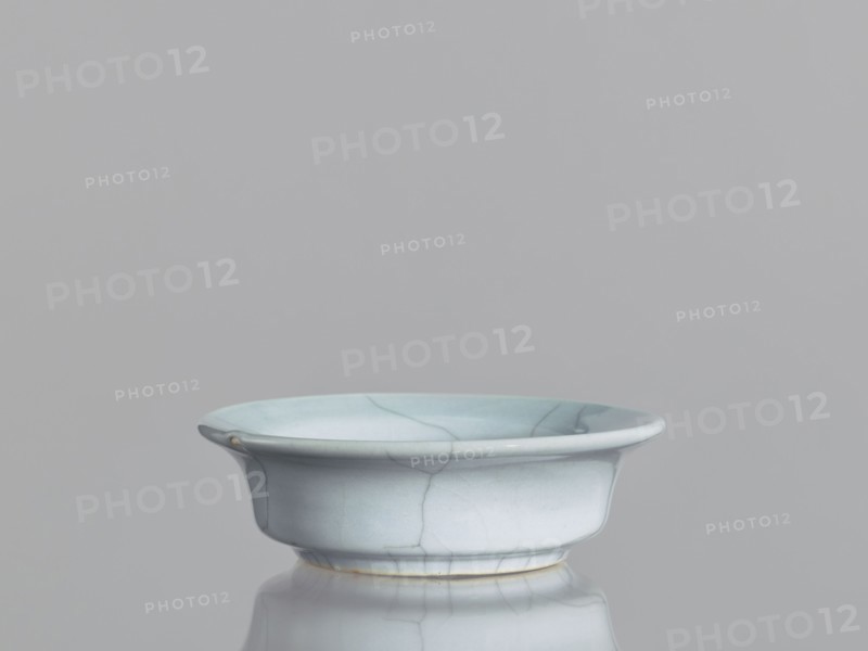 Flowerpot saucer, Chinese ceramics, Qing 1644–1911, 19th century. Grey-blue glaze with cracklework; glazed ceramic vessel, saucer, height 3.5 cm, diameter 11.4 cm., A shallow bowl with a flared rim sits on a reflective surface, showing a smooth, glazed finish with visible crackle lines; the bowl is primarily pale blue with subtle variations of light blue and hints of white, and there are small areas of warm beige near the base and a tiny spot on the rim, all against a uniform gray background.