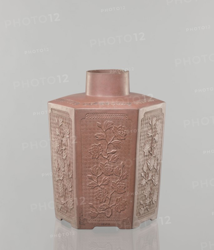 Yixing clay storage vessel; maker, red-brown stoneware with plastic, impressed decoration; decorated with peonies; Qing dynasty 1644–1911, 19th century; Yixing style; Chinese; vessel; height 22.4 cm, width 17.7 cm, depth 15.7 cm, weight 1.15 kg., A hexagonal ceramic vessel with a short cylindrical neck, shown against a pale gray background. The vessel is predominantly reddish-brown with lighter beige accents along the edges of its panels. Each of the visible vertical panels features raised floral reliefs with petals and leaves, rendered in the same reddish-brown and lighter beige tones. The top surface around the neck shows a subtle decorative band in the reddish-brown color. The base has small notches and rests on a reflective surface that mirrors the colors and shapes faintly.