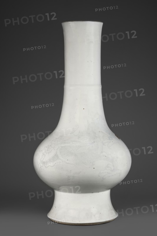 Vase, Chinese, Qing dynasty 1644–1911, 19th century. Porcelain body, spherical, flattened horizontally on a high foot; high, wide neck with a rim. Decoration: dragons engraved under the glaze into the body and filled with cobalt. Height 42.7 cm; diameter 21.5 cm., A tall bulbous ceramic vase with a long cylindrical neck and a flared foot, rendered in white with very faint pale gray decorative motifs visible on the rounded body, sitting against a dark gray background; the vase surface shows a few small dark specks and subtle irregularities in glaze, and the rim and base edges reveal a thin darker brownish-gray line.