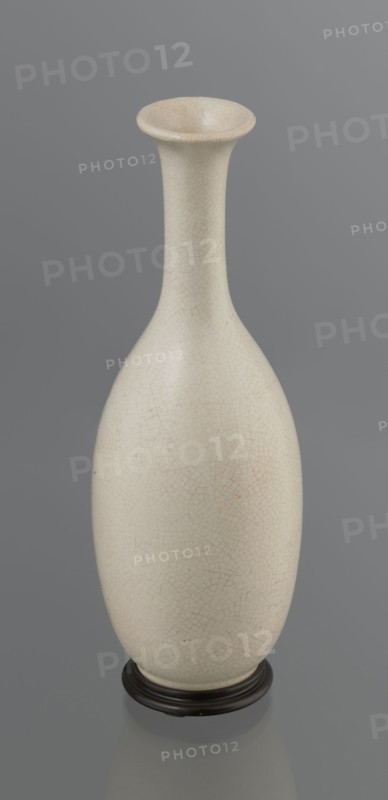 Vase on saucer. Vase made of stoneware or thick porcelain?, ovoid with a long cylindrical neck flaring at the spout; light cream white matte glaze covered with fine cracklework. 19th century, Japan, Edo 1603–1868. Ceramics, bottles. Height 24.1 cm, diameter 8.6 cm, weight 0.65 kg., A tall, narrow-necked vase with an elongated bulbous body stands on a low round base; the vase surface shows a fine network of hairline crackle patterns and is an off-white or cream color with subtle warm beige variations, while the base appears dark brown to nearly black. The background is a smooth, muted gray.