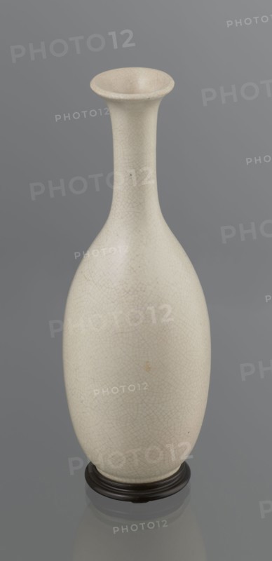 Vase on saucer. Vase made of stoneware or thick porcelain?, ovoid with a long cylindrical neck, flaring at the spout. Light cream white matte glaze covered with fine cracklework. 19th century, Japan, Edo 1603–1868. Ceramics; cracklework decoration; bottles. Vessel. Height 24.1 cm, diameter 8.6 cm, weight 0.65 kg., A tall, narrow-necked ceramic vase with a flared rim stands on a dark round base; the vase surface is a pale off-white or cream with a fine crackle pattern and a few small darker specks and slight discolorations, and the background is a uniform mid-gray.