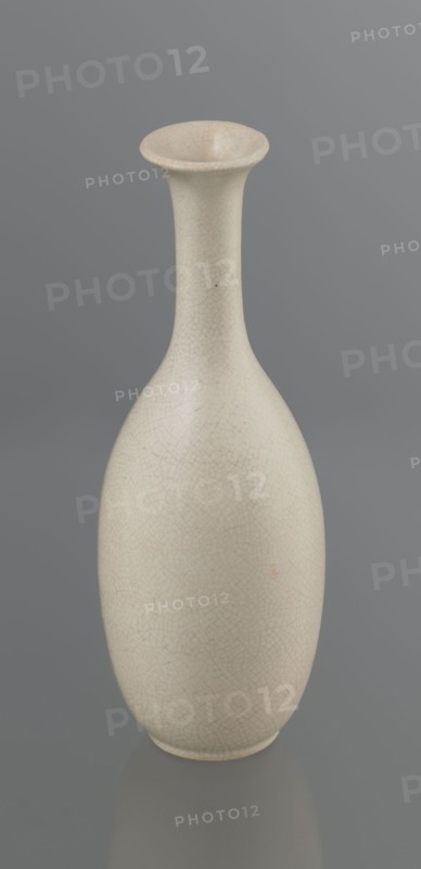 Vase on saucer. Vase made of stoneware or thick porcelain?, ovoid with a long cylindrical neck flaring at the spout. Light cream white matte glaze covered with fine cracklework. 19th century, Japan, Edo 1603–1868. Ceramics; bottles. Height 24.1 cm, diameter 8.6 cm, weight 0.65 kg., A tall, slender vase with a narrow neck that flares at the rim sits against a smooth gray background; the vase surface is an off-white or cream color with a fine network of subtle crackle lines and slight variations in shading, and there are small darker specks and a faint warm-toned spot on the body.