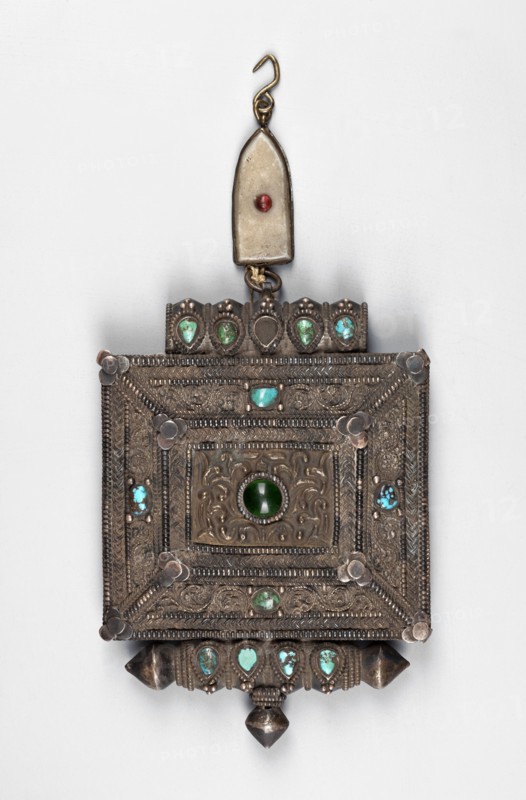 Ga'u box, gift, Himalayan Tibetan Buddhist amulet/ceremonial container, 19th/20th century, Tibet; materials: jewelry/amulet; height 19 cm, width 8.5 cm, depth 2.3 cm., A rectangular metallic pendant with ornate engraved patterns and raised borders, featuring decorative round and teardrop cabochon stones in shades of green and turquoise set into the metal; a central round green stone is surrounded by embossed floral-like motifs, with several small turquoise-green stones arranged along the top and bottom edges and at corners; the metal shows a patinated silver-gray and bronze tone, with darker shadowed areas and small highlights; above the main pendant a small elongated metal plate with a single small round red bead is attached to a hooked suspender in a brass-like color.