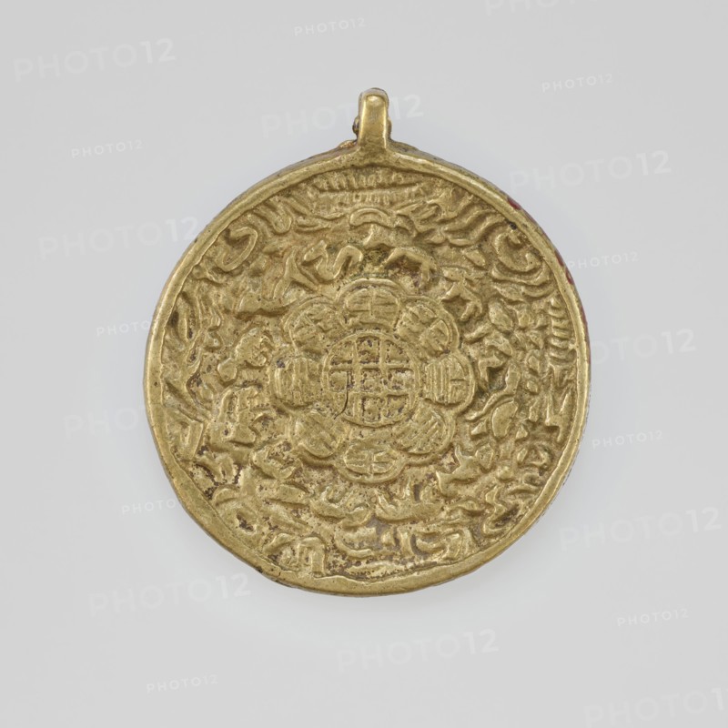 Amulet, 19th–20th century, Tibet; Tibetan Buddhist art; amulet with figural decorations; material/technique: jug-like form; height 5.9 cm, diameter 5.2 cm., A circular metallic pendant with a small loop at the top, primarily gold in color, showing a raised decorative pattern across its surface that includes a central floral-like motif surrounded by concentric ornamental designs and textured relief; the background around the pendant is a light gray.