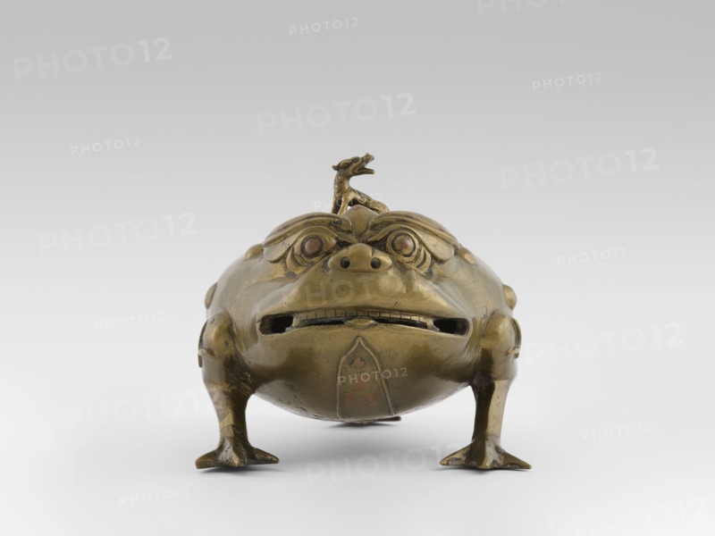 Incense burner with a cover — moon frog toad. Cast in bronze studded with copper. Popular motif in Chinese geomancy — symbol of a three-legged frog toad bringing money wealth. 19th century, Burma Myanmar. Burmese art; ceremonial vessel; incense burner. Motifs: frogs, coins, tiger; symbols of wealth and luck. Height 20.0 cm; width 32.0 cm; depth 21.0 cm., A metallic sculptural object shaped like a squat creature with a rounded body, short legs, and a wide mouth is shown from the front; the surface appears largely brass-colored with areas of darker patina and hints of reddish-brown on the eyes and some worn spots. The creature has stylized facial features including prominent brows, circular eyes set into carved sockets, a small raised nose area with visible nostril openings, and a horizontal mouth opening with a suggestion of teeth. A small ornamental figure sits atop the creature’s head. The legs end in flattened, webbed-looking feet that rest on a pale, neutral background that transiti...