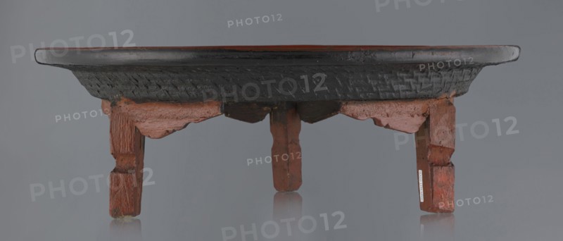 Ritual vessel, gift; Burmese art, Burmese culture, religious rites. Tray with a fluted surface showing a bamboo weaving pattern, made of bamboo covered with black lacquer on the bottom. Profiled legs covered with red lacquer. Overall good condition; local abrasions and cracks in the lacquer layers, many local cracks and lacquer losses on the bottom. 19th/20th century, Burma Myanmar. Vessel; height 15.5 cm, diameter 44.5 cm., A low wooden table seen from the front with a round dark tabletop and a carved dark band underneath, supported by a group of short carved legs and decorative aprons in a reddish-brown wood tone; the surface shows areas of worn dark finish and the wood grain and carving details are visible, with the background and floor appearing in muted gray and reflections of the legs faintly visible on the surface beneath.