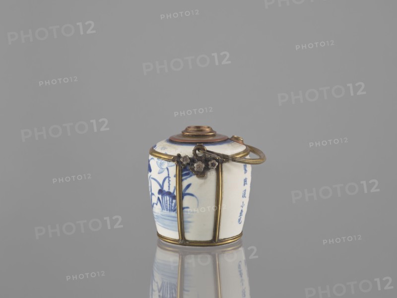 Water pipe, Porcelain belly mounted in a metal frame with floral ornament. Porcelain painted underglaze with cobalt matte glaze. 19th century. Decoration: lotuses, ducks; inscriptions in Chinese. Materials/technique: ceramics, blue-and-white porcelain. Function: pipe/smoking accessory. Dimensions: height 11.0 cm, width 14.6 cm, depth 10.1 cm. Cultural attributions: Southeast Asia Vietnamese?, China., A small ceramic container with a rounded lid sits centered on a reflective surface against a plain gray background; the container is primarily white with blue painted floral and plant motifs on its visible sides, and it is framed with brass-colored metal bands and fittings including a decorative clasp and a curved handle; the lid has brownish and blue accents, and the reflection of the object appears below it.