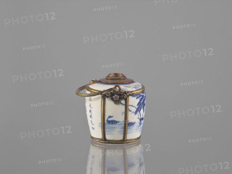 Water pipe, Porcelain belly mounted in a metal frame with floral ornament. Porcelain painted underglaze with cobalt matte glaze. 19th century. Possibly Vietnam or China. Blue and white porcelain. Depicts lotuses and ducks. Smoking accessory; pipe. Inscriptions present. Height 11.0 cm, width 14.6 cm, depth 10.1 cm. Cultures referenced: Southeast Asia, Vietnamese, Chinese., A small cylindrical ceramic container with a rounded lid sits on a reflective surface, showing a porcelain body decorated with blue floral and bird motifs and a few areas of blue brushwork, encased in a brass or gold-toned metal frame with vertical bands and a hinged decorative clasp, and a metal loop handle attached near the top; visible colors include white, blue, gold, brown, and gray in the background and reflection.