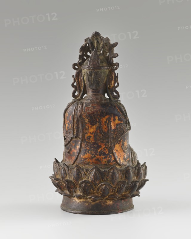 Guanyin; creator, Chinese; Qing dynasty 1644–1911, 18th century; sculpture; material not specified; height 24 cm; subject: bodhisattva Guanyin seated on a lotus throne; iconography: mercy; Buddhist art; Chinese Buddhism; figurine., A rear view of a seated figure carved from dark material with worn areas showing mottled patches of orange and brown, the figure wearing an ornate crown or headdress with swirling carved details and hair rendered in vertical striations, shoulders draped in a flowing garment with textured folds, and the figure seated on a stylized lotus base with layered petal shapes in dark brown and hints of gold and reddish wear against a pale gray gradient background.