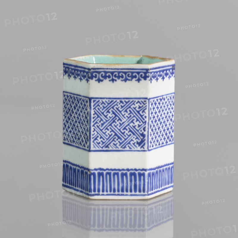 Bowl holder, Hexagonal prism-shaped container for incense or bowls, decorated with three stripes of geometric borders and meander ornament. Porcelain body with white exterior glaze, overglaze decoration with cobalt enamel; interior and foot glazed in turquoise; bronze foot and rim edge. Signed. Chinese, Qianlong style Qing dynasty, 1644–1911, 19th century . Height 7.5 cm, width 5.8 cm., A hexagonal ceramic vessel sits on a reflective surface against a gray background, showing white and blue exterior glazing with a pale turquoise interior rim; the sides feature decorative bands including a top scrolling motif in blue, a central section with geometric and lattice-like patterns in blue on white, and a lower band of vertical blue strokes above the base, with subtle signs of wear along the rim and a soft reflection beneath.