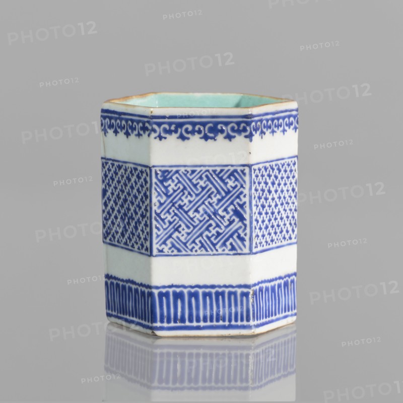 Hexagonal bowl holder in the shape of a prism, decorated with three bands of geometric borders and meander ornament. Porcelain body with white glaze outside; overglaze decoration with cobalt enamel; interior and foot glazed in turquoise; bronze foot and rim edge. Signed. Incense container, colored porcelain, Qing dynasty Qianlong style, 18th–19th century?, Height 7.5 cm, width 5.8 cm., A hexagonal ceramic vessel stands on a reflective surface against a pale gray background, showing a white body with decorative bands of deep blue patterns and a pale green interior; the top rim has a thin brown edge, the upper band features a scrolling blue border on white, the central band displays alternating intricate geometric blue motifs on white, and the lower band has vertical blue fluting on white, with a faint reflection visible beneath.
