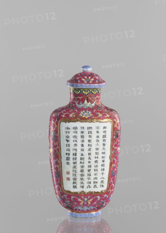 Vase with lid, Chinese, Qing dynasty Qianlong period, 1736–1795. Flattened vase form; colored porcelain with lid; lotus tendril ornament; inscribed poems/poetry. Dimensions: height 22.7 cm, width 10.7 cm, weight 0.65 kg., A slender, lidded vase with a rounded shoulder and narrow neck stands against a plain light gray background; its surface is predominantly deep red with ornate floral and scrolling patterns in colors including yellow, blue, green, white, pink, and gold; the lid and base feature bands with geometric motifs in blue and white, and the lid has a small white knob topped with blue decoration; a recessed rectangular panel on the front shows a white area framed in gold surrounded by the red patterned surface; the overall form is glossy and reflective, and the image shows the vase upright with a faint reflection visible below it.