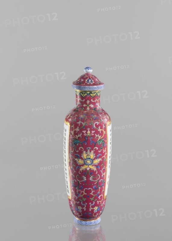 Vase with lid; Chinese; Qing dynasty, Qianlong period 1736–1795; colored porcelain; flattened vase form with lid; lotus tendril ornament; inscribed with poems/poetry; height 22.7 cm, width 10.7 cm, weight 0.65 kg., A tall, slender ceramic bottle with a domed lid stands against a plain light gray background; the bottle is predominantly deep red with ornate scrolling floral and foliate patterns in colors including yellow, blue, green, white, and pink, and it has decorative bands near the neck and base in blue and white; the lid echoes the red and multicolored floral motifs and is topped with a small rounded knob that shows blue and white; the bottle shows two vertical panels of a lighter color on opposite sides, each framed by a thin gold border, and the surface has a glossy finish that reflects light.