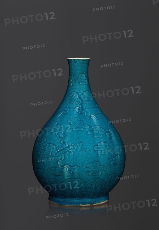 Vase in the shape of an onion-shaped bottle, Chinese art, Chinese culture, Qing 1644–1911 style, hundred antiquities baigu ornament, ceramics. Onion-shaped bottle-vase with a narrow, short neck. The vessel is decorated with a flat relief creating a composition combining a cloud and babao emblems eight precious objects or eight basic symbols. Glaze is single-color, dark, in green-blue tones. 16th–17th century, China. Vessel; height 27 cm; weight 2.6 kg., A teardrop-shaped ceramic vase with a narrow neck and flared rim sits against a dark gray background, the vase surface showing a vibrant turquoise-blue glaze with slightly darker and lighter variations, decorated with an all-over raised pattern of stylized cloud motifs and flowing ribbon-like elements, and a band of repeating wave-like patterns near the base; the rim and foot show a thin exposed edge of unglazed clay in a light beige color, and the vase casts a subtle reflection on the surface beneath it.