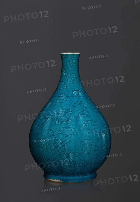 Vase in the shape of an onion-shaped bottle, Chinese, Qing 1644–1911, 16th–17th century, ceramics. Onion-shaped bottle-vase with a narrow, short neck, height 27 cm, weight 2.6 kg. Decorated in flat relief with a composition combining cloud and babao eight precious objects emblems. Single-color dark glaze in green-blue tones., A teardrop-shaped ceramic vase in a vivid turquoise-blue with a narrow neck and flared rim standing against a dark gray background; the vase surface shows an embossed or incised pattern of stylized cloud-like motifs and flowing ribbon shapes, with a band of wave-like decorative patterns near the rounded base, and a small area of exposed lighter clay where the glaze is worn at the bottom edge.